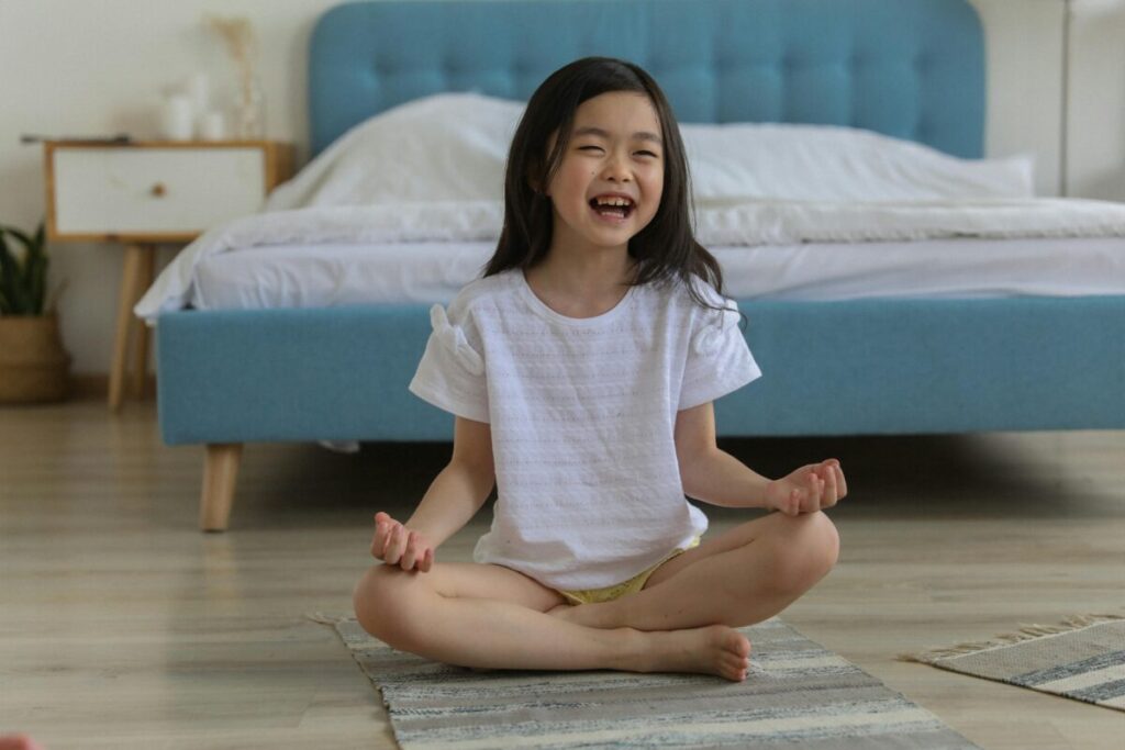 A cheerful young girl practicing yoga indoors, seated cross-legged and smiling brightly in a cozy bedroom.